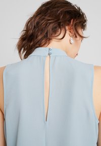 Blusa senza maniche azzurro chiaro con colletto alto, dotata di un'apertura a forma di chiave sulla schiena e due piccoli bottoni per la chiusura. Tessuto leggero e morbido.