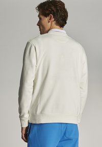 Svetlo kremast pulover z dolgimi rokavi, okroglo ovratnikom in rebrastim robom. Zdi se mehak z gladko teksturo in minimalističnim dizajnom.