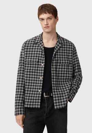 Black and white checkered shirt made of cotton with a collar, front buttons, and two chest pockets. Worn over a black t-shirt.