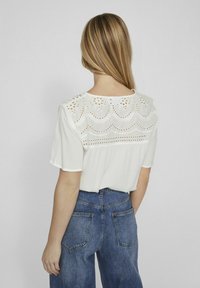 Blouse blanche à manches courtes avec un empiècement brodé détaillé et des motifs ajourés, associée à un jean en denim bleu taille haute.