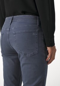 Personne portant un jean en denim bleu et un t-shirt noir à manches longues vu de dos, mettant l’accent sur la poche et les détails de couture du jean.