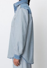 Valkoinen denim-paita, jossa on napillinen kaulus, pitkät hihat ja rintatasku. Siinä on hieman kaareva helma ja kontrastiväriset hihansuut.