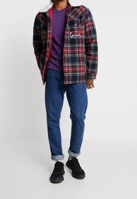 Rutigt jacka med mönster i rött, marinblått och gult, vit konstpäls-kant, framfickor, lager över en lila skjorta och blå jeans. Svarta skor.