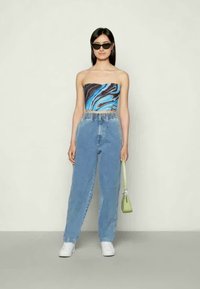 Top sem alças com um padrão em mármore azul e preto, combinado com jeans de cintura alta azul claro e sapatilhas brancas. Segura uma pequena mala verde.