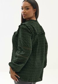 Blouse vert foncé texturée avec des accents froncés et des détails à motifs sur les manches, offrant une coupe décontractée et des manches longues.