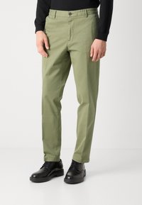 Uomo che indossa pantaloni dritti di un verde oliva chiaro, una maglietta nera a maniche lunghe e scarpe stringate nere su uno sfondo bianco.