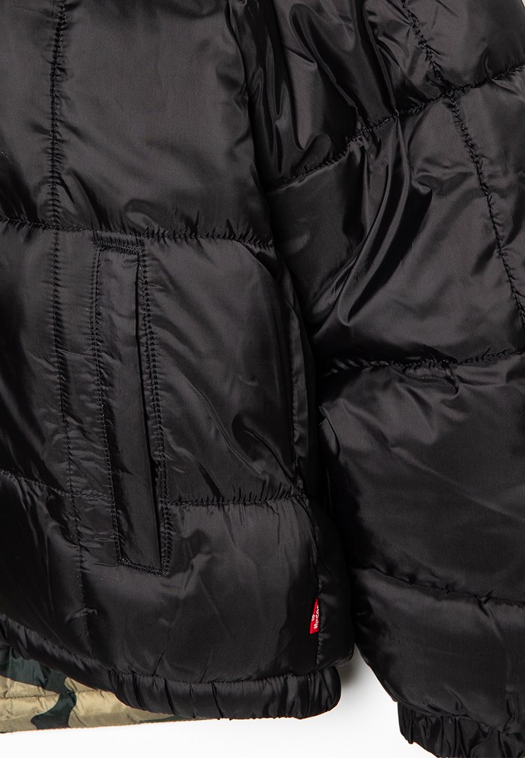 Veste doudoune noire avec une texture lisse et brillante. Présente des coutures horizontales, des poignets élastiques et une poche latérale. Bordée de tissu camouflage.