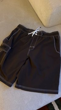 Schwarze Badeshorts mit weißer Kontrastnaht, weißem Kordelzug an der Taille und einer Seitentasche mit schwarzem Text auf einer beigen Stoffoberfläche.