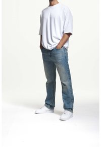 Valkoinen ylisuuri t-paita, joka on yhdistetty vaaleansinisiin farkkuihin ja valkoisiin lenkkareihin. Kangas näyttää pehmeältä, siinä on rento leikkaus ja kevyttä kulumista.