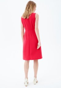 Vestido sin mangas rojo con un corsé ajustado, dobladillo acampanado y detalles en las costuras. Combinado con tacones metálicos de bloque. El fondo es blanco puro.