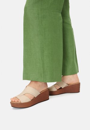 Beżowe sandały na koturnie z dwoma szerokimi paskami, drewnianą platformą i fakturowaną podeszwą, zestawione z zielonymi spodniami o szerokich nogawkach.