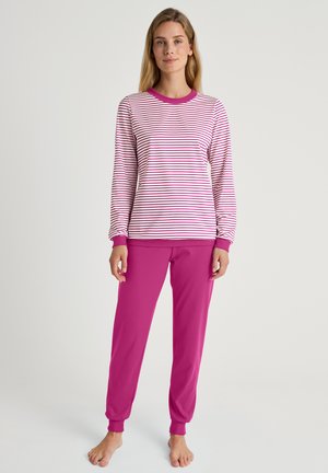 Vrouw staat op blote voeten, draagt een witte lange mouw shirt met roze strepen en effen roze broek tegen een effen lichte achtergrond.