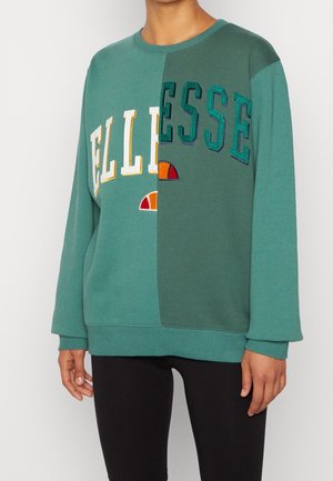 Henkilö pukeutunut vihreän sävyiseen väriblokkipaiseen collegepaitaan, jossa on "ELLESSE"-teksti ja logo keskeltä jaettuina, yhdistettynä mustiin housuihin.