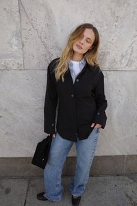 Must黑色 särg valge triibulise särgi peal, kergete siniste laiade teksadega, mustade lapikute kingade ja musta koti kõrval kiviseina taustal.