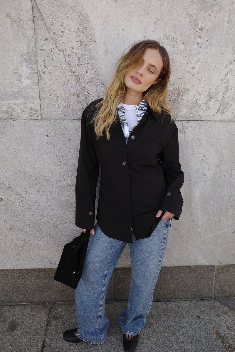 Must黑色 särg valge triibulise särgi peal, kergete siniste laiade teksadega, mustade lapikute kingade ja musta koti kõrval kiviseina taustal.