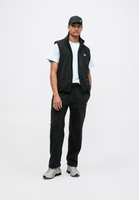 Vesta din fleece negru peste un tricou albastre deschis, asortată cu pantaloni negri și pantofi sport gri. Prezintă un fermoar și accente cu logo-ul Nike.