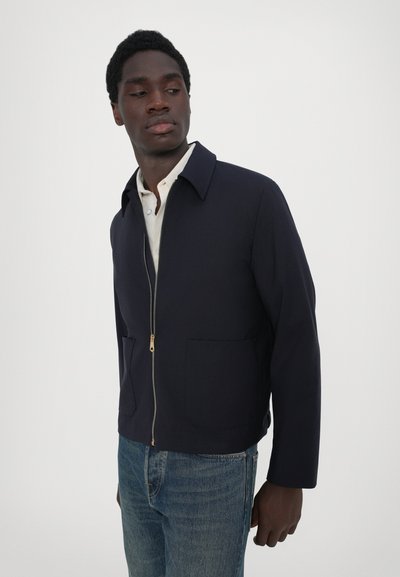 Giacca blu navy con zip e colletto a punta, due tasche frontali e dettagli della zip dorati, abbinata a jeans azzurri. Texture liscia.