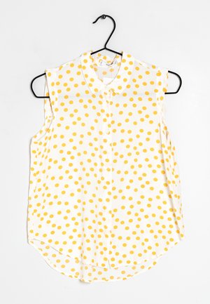 Blouse blanche sans manches à pois jaunes, fermeture boutonnée sur le devant et col, suspendue à un cintre noir.