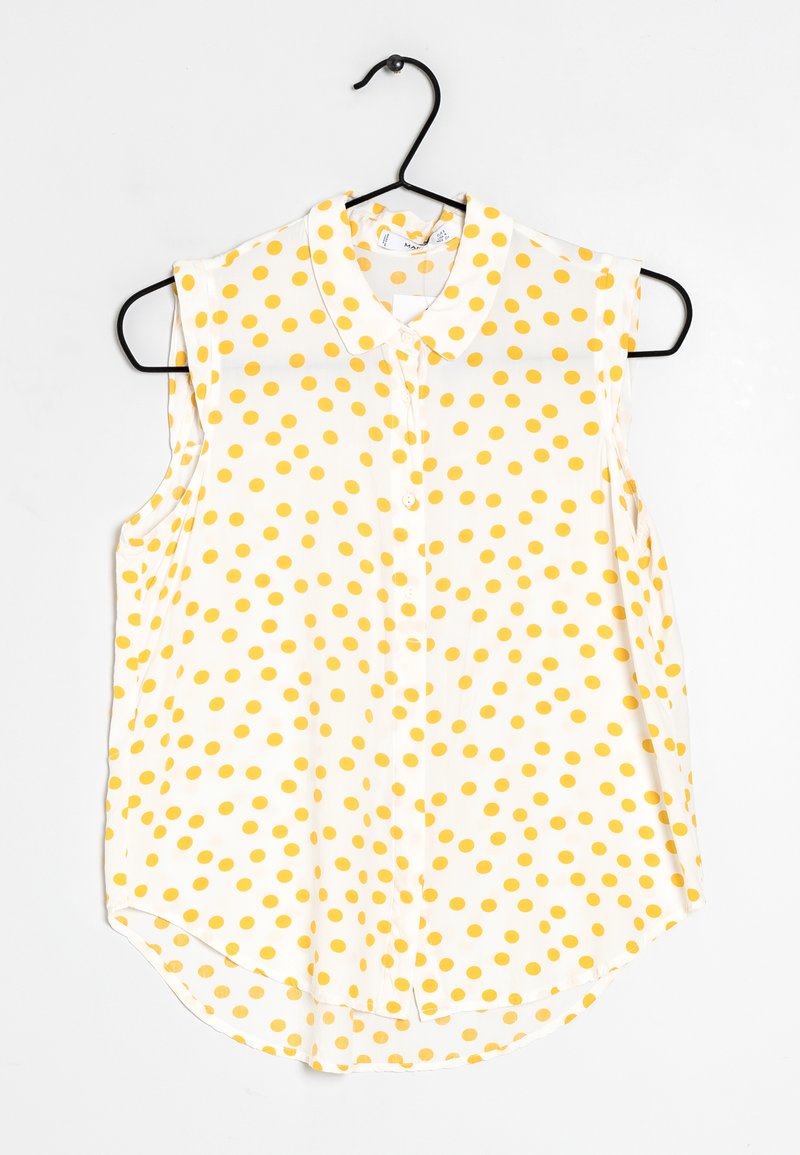 Blouse blanche sans manches à pois jaunes, fermeture boutonnée sur le devant et col, suspendue à un cintre noir.