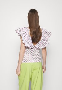 Top bianco con stampa floreale rosa, caratterizzato da ampie maniche arricciate e una profonda scollatura sulla schiena, abbinato a pantaloni larghi verde chiaro.