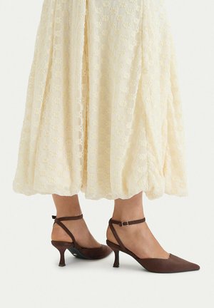 Mujer vistiendo una falda midi texturizada color crema y zapatos slingback marrones de ante con tacón bajo, punta puntiaguda y tiras en el tobillo.