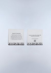 Dos etiquetas blancas de prendas con el logo negro de Napapijri en la parte inferior; una detalla un tratamiento especial de tintura, la otra indica el uso del 70% de materiales reciclados.