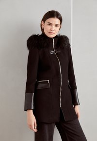 Cappotto nero con cappuccio in pelliccia sintetica, zip metallica e dettagli in pelle sui polsini e sulle tasche. Vestibilità sartoriale con un design elegante.