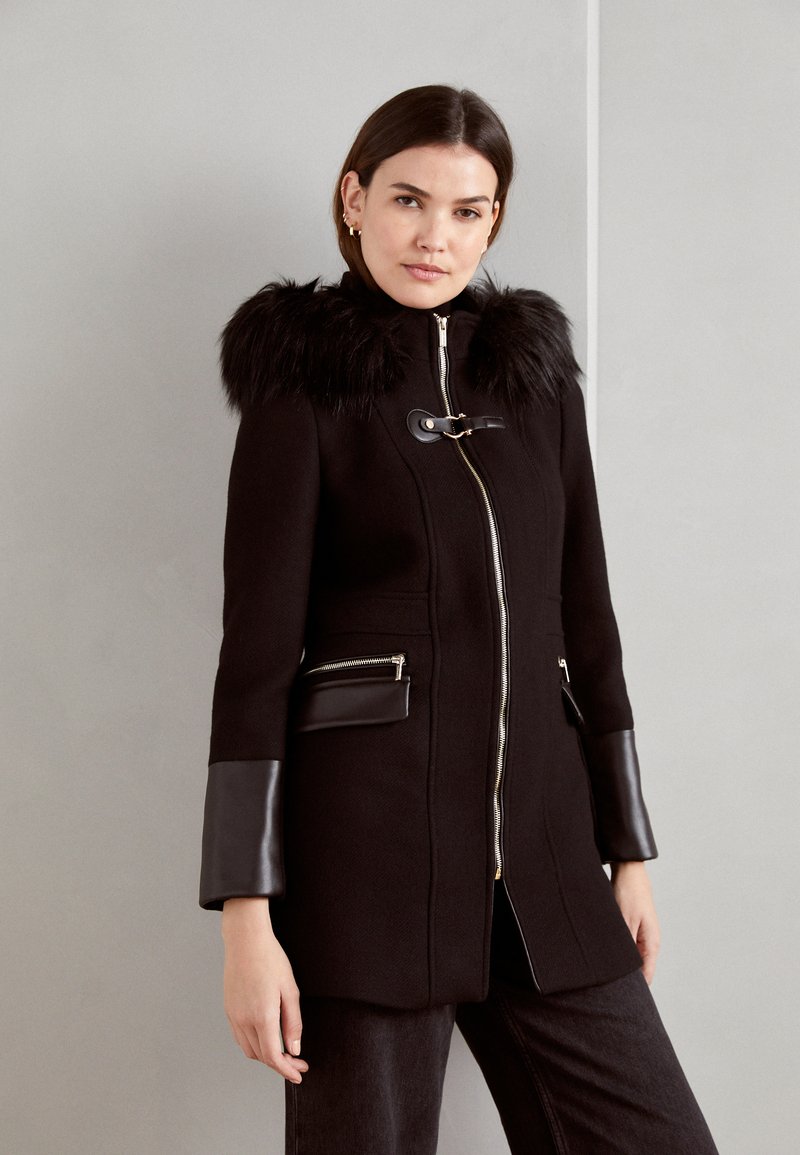 Cappotto nero con cappuccio in pelliccia sintetica, zip metallica e dettagli in pelle sui polsini e sulle tasche. Vestibilità sartoriale con un design elegante.