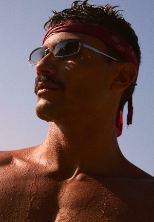 Uomo con baffi e pelle bagnata che indossa occhiali da sole e una bandana rossa, guarda di lato contro un cielo azzurro limpido.