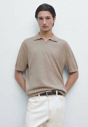 Jonge man met donker haar, gekleed in een beige korte mouw polo, witte broek en een gevlochten zwarte riem, staand tegen een witte muur.