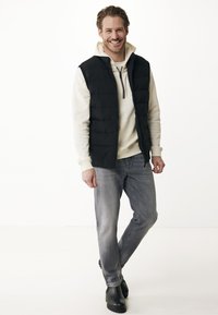 Muž stojící a usmívající se, oblečený v černé vycpané vestě přes světle šedou mikinu, šedých džínách a černých botách na bílém pozadí.