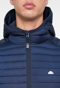 Meriinsinine puffer-jope tõmblukuga esiküljel ja kapuutsiga. Omadusteks tikitud muster, sile tekstuur ja väike valge logo rinnal.