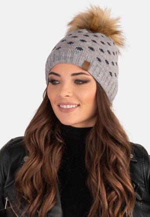 Bonnet en tricot gris avec des pois noirs, surmonté d'un pompon en fausse fourrure. Présente une petite étiquette en cuir marron sur le côté.