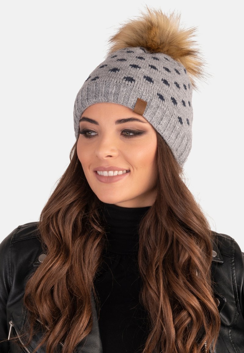 Grå strikket hue med mørke prikker, toppet med en faux fur pom-pom. Har en lille brun læderlabel på siden.