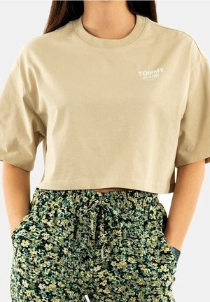 T-shirt court beige Tommy Jeans associé à un pantalon taille haute à cordon, à motif floral vert et jaune, mains dans les poches.