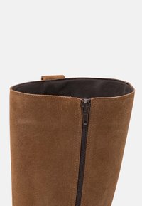 Brązowy zamszowy kozak do kolan o gładkiej fakturze, z bocznym zamkiem błyskawicznym i pętelką u góry, ułatwiającą zakładanie.