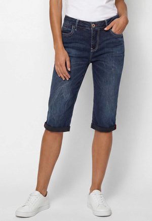 Person trägt hochgekrempelte dunkelblaue Denim-Knielange Shorts, weiße Sneakers und ein weißes Oberteil und steht vor einem einfarbigen Hintergrund.