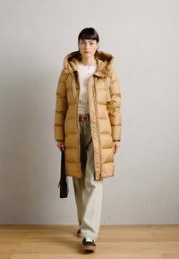 Beige Daunenjacke mit Kapuze, gestepptem Stoff, vorderem Reißverschluss und Seitentaschen, kombiniert mit hellen Hosen und braunen Schuhen.