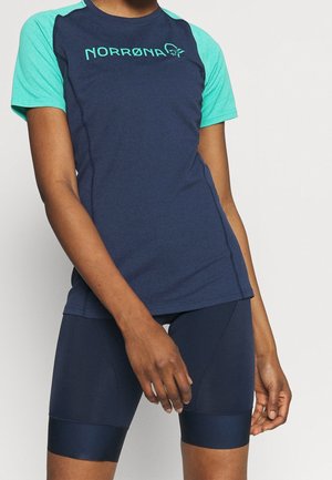 Person trägt ein marineblau-türkisfarbenes sportliches Kurzarmshirt mit dem "NORRØNA"-Logo und eng anliegende marineblaue Radhosen.