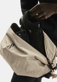 Ręka wkłada czarne buty sportowe do otwartej beżowej torby na zamek błyskawiczny, z widocznym logotypem marki.