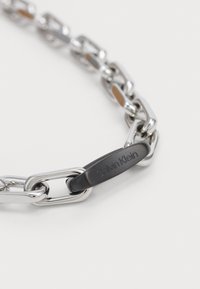 Hopeaketju, jossa on toisiinsa kytkeytyviä renkaita. Musta riipus, jota koristaa "Calvin Klein" -kaiverrus. Sileä metallinen pinta.