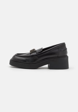 Mocassin en cuir noir avec un talon bloc épais et une boucle décorative sur le dessus, vu de côté sur un fond uni.