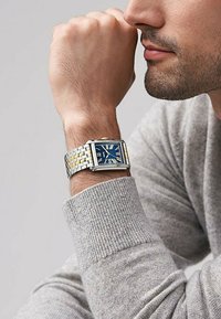 Montre en métal argenté et doré avec un cadran rectangulaire. Le cadran bleu présente des chiffres romains. Portée au poignet, un pull gris texturé est visible.