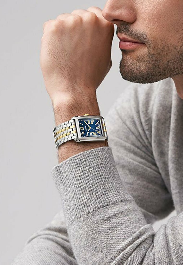 Montre en métal argenté et doré avec un cadran rectangulaire. Le cadran bleu présente des chiffres romains. Portée au poignet, un pull gris texturé est visible.