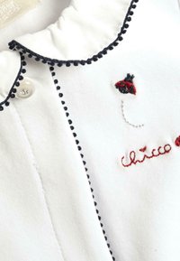 Camicia di cotone bianca con colletto arricciato, bordi blu navy, coccinella ricamata e testo "chicco" in rosso. Presenta una chiusura a bottone.