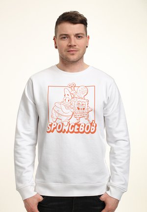 Mężczyzna w białej bluzie z pomarańczowym rysunkiem linii postaci SpongeBoba, Patricka i Gary’ego, uśmiechających się, z rękami w kieszeniach, skierowany przodem.