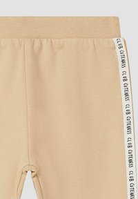 Beige sweatpants met een gestructureerde tailleband, uitgerust met een zijstreep waarin "CLUB CUTENESS" in het zwart is gedrukt. Glad materiaal met stikseldetails.