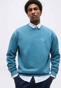 Jonge man met handen in de zakken, gekleed in een teal crewneck-trui over een wit en lichtblauw gestreept overhemd met knopen.