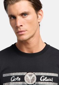 Zwarte katoenen sweatshirt met een ronde halslijn, wit Grieks sleutelpatroon en een centraal zilveren logo met een gevleugeld figuur en merknamen.