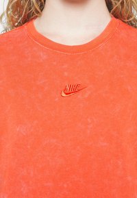 Oranžové bavlněné tričko s krátkým rukávem a kulatým výstřihem, na levé části hrudi má malé vyšívané logo Nike v červené barvě. Viditelná jemná textura.